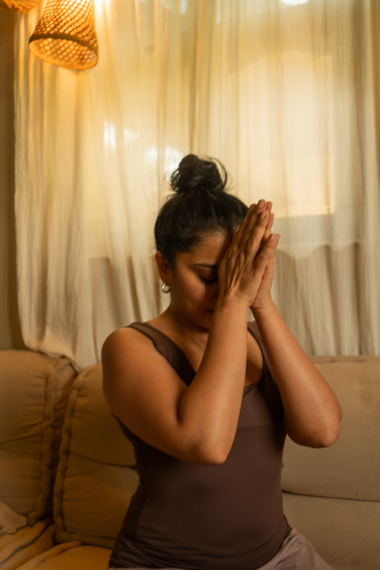 Alisha Netalkar seated in meditation with hands held in prayer at her forehead, in a warmly lit room